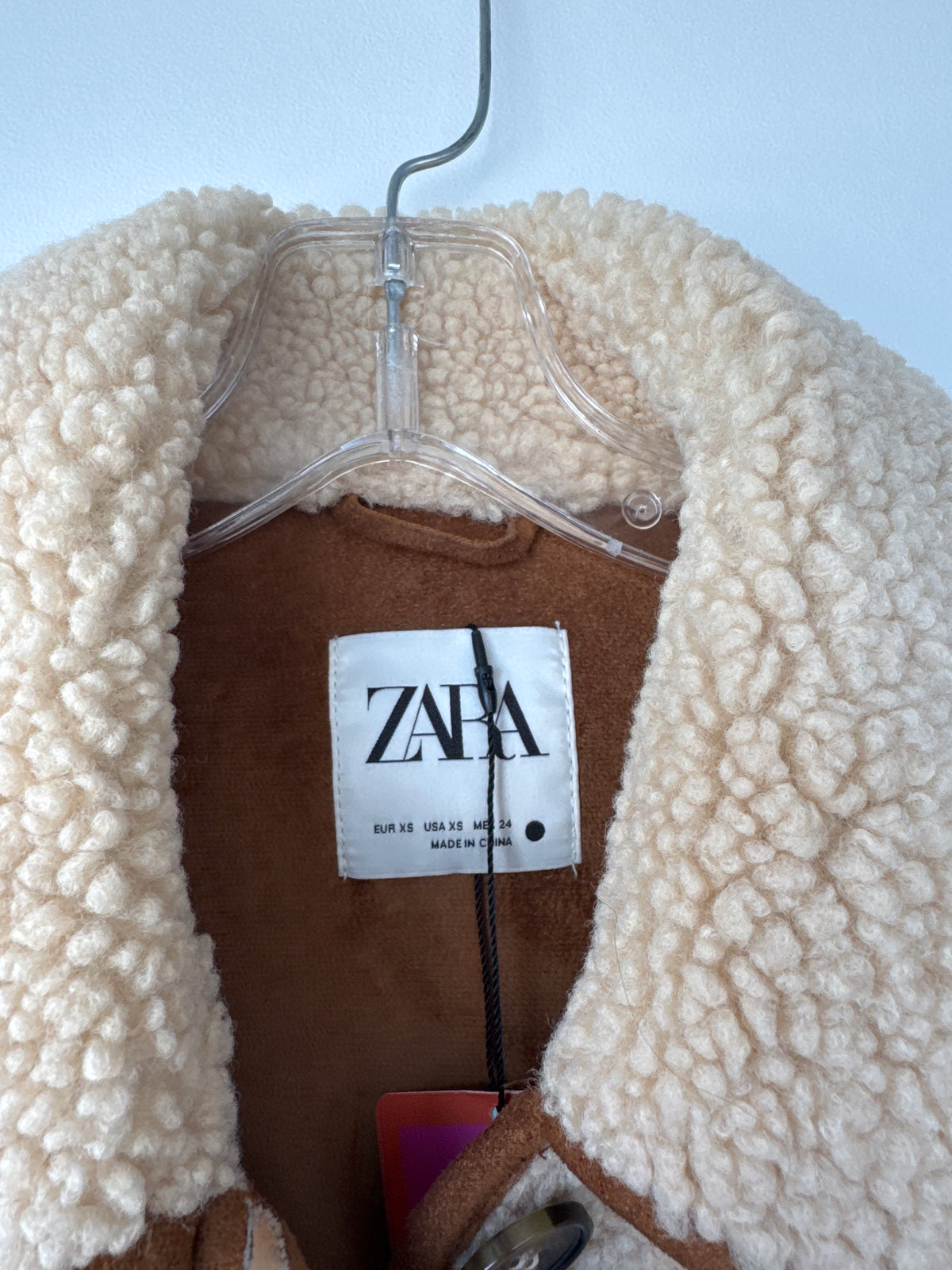 A close-up of a ZARA shearling jacket hanging on a clear plastic hanger against a light blue background. The jacket features a thick, textured cream-colored sherpa collar and a brown inner lining, with the white ZARA brand label clearly visible in the center. A beige sherpa fleece button-up coat with brown trim and a brown inner lining, hanging on a metal hanger against a light blue wall. Zara teddy bear jacket, Zara teddy jacket, women’s teddy jacket, teddy fleece jacket, sherpa jacket, button-down jacket,