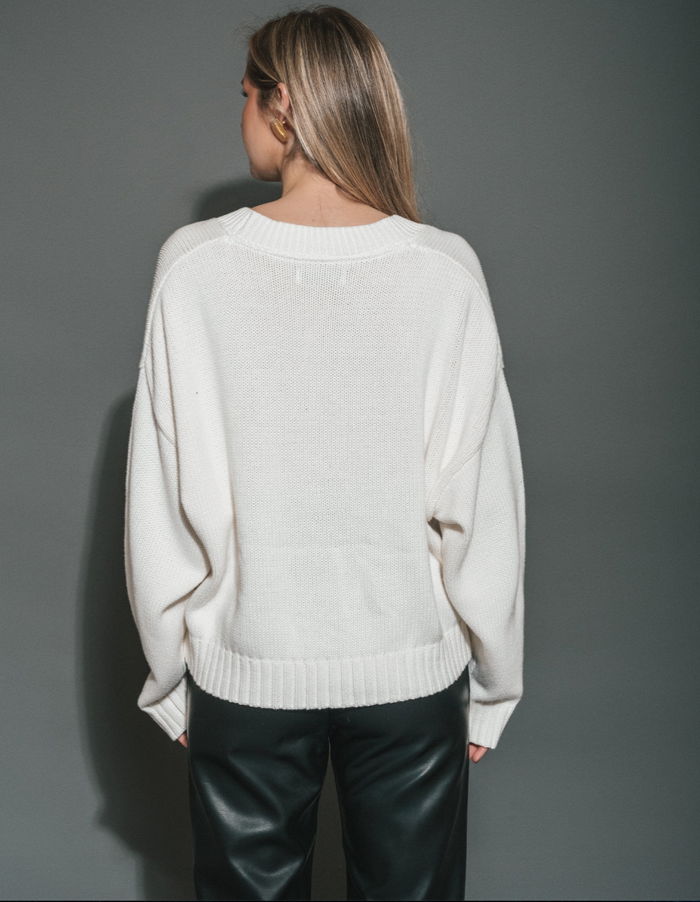 A woman viewed from behind wearing an oversized off-white knit sweater and black leather pants against a dark gray studio background. SHPK consignment, Sherwood Park shop local. Brunette The Label cheaper