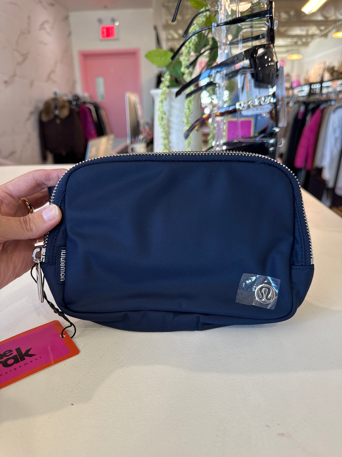 A Lululemon Belt Bag navy blue nylon pouch bag with a silver zipper and a small silver logo patch on the front, held by a hand in a retail store.