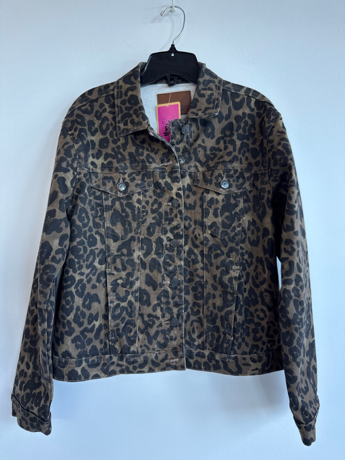 A leopard print denim jacket hanging on a black hanger against a plain light gray wall. The jacket features a brown and black animal print pattern with a button-up front and two chest pockets. Judy Blue. Sherwood Park consignment, Sherwood Park shopping, consignment store Sherwood Park, Edmonton consignment, secondhand shopping, secondhand clothing, women’s consignment, women’s clothing consignment, curated consignment, quality secondhand clothing, pre-loved clothing, thrift shopping, sustainable fashion, l