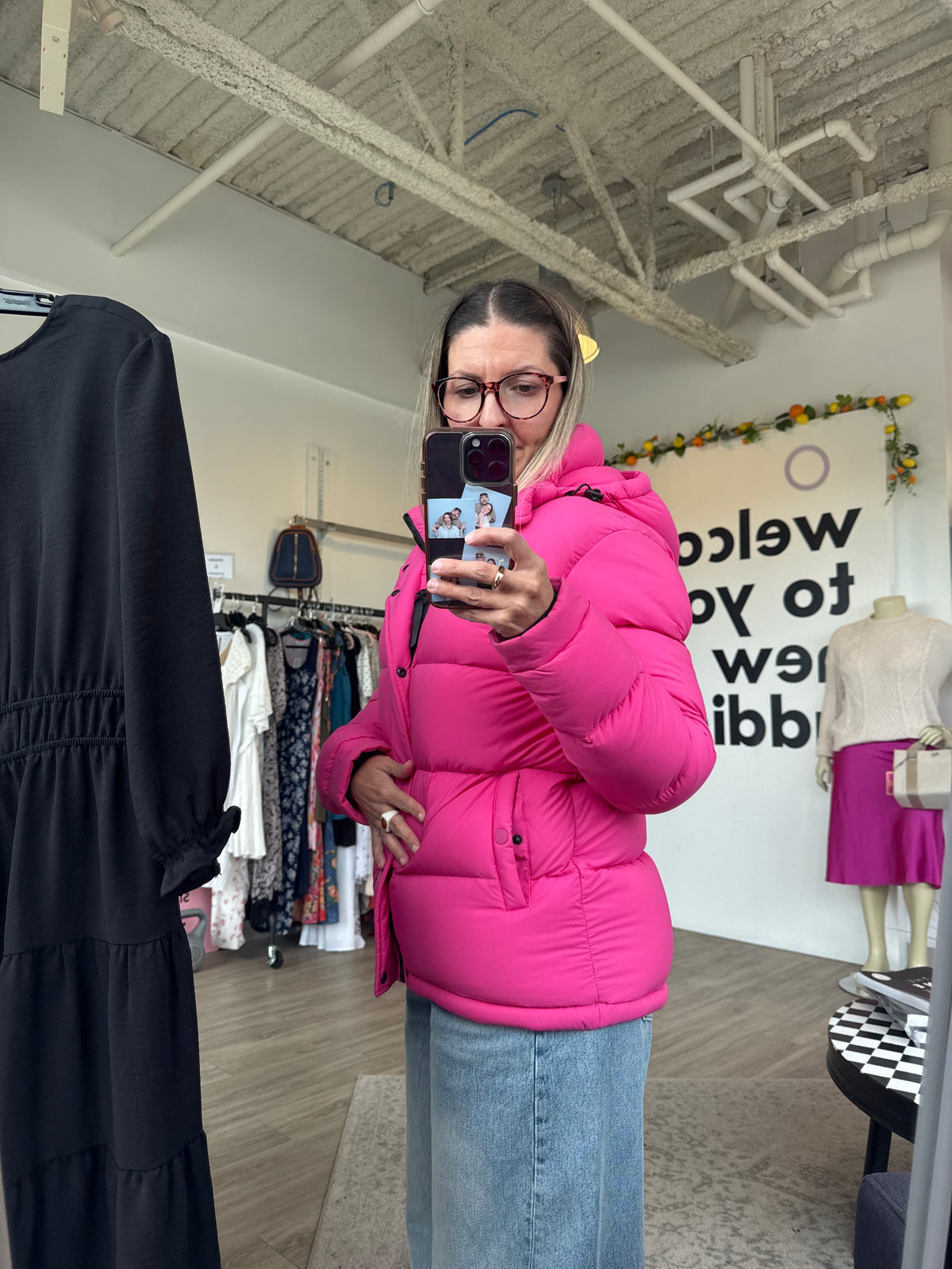A woman takes a mirror selfie in a boutique while wearing a bright pink puffer jacket and a light blue denim skirt. The boutique has a rustic industrial ceiling with exposed beams, clothing racks, and a mannequin in the background. Aritzia Super Puff, Super Puff jacket, Aritzia puffer jacket, Aritzia winter jacket, women’s puffer jacket, women’s winter coat, down puffer jacket, insulated jacket, warm winter jacket, hooded puffer jacket, Aritzia coat, winter outerwear, cold weather jacket, Canadian winter ja
