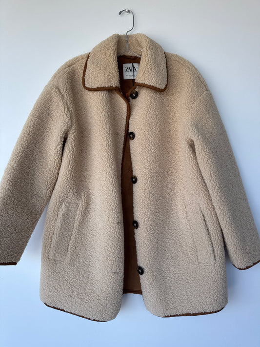 A beige sherpa fleece button-up coat with brown trim and a brown inner lining, hanging on a metal hanger against a light blue wall. Zara teddy bear jacket, Zara teddy jacket, women’s teddy jacket, teddy fleece jacket, sherpa jacket, button-down jacket, cozy women’s jacket, plush jacket, fall jacket, winter jacket, Edmonton consignment, women’s clothing consignment