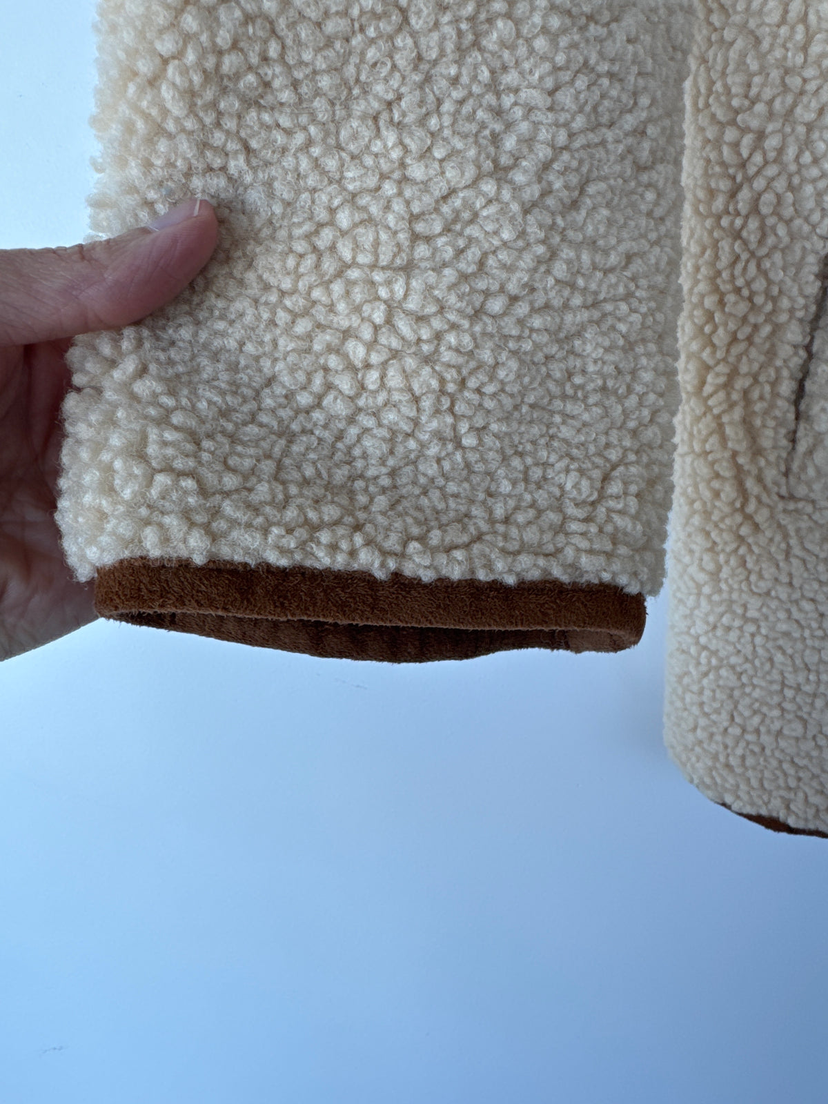 A close-up of a cream-colored sherpa fleece garment being held by a hand, showing the soft textured fabric and a contrasting brown suede-like trim at the cuff, against a light blue background.
A beige sherpa fleece button-up coat with brown trim and a brown inner lining, hanging on a metal hanger against a light blue wall. Zara teddy bear jacket, Zara teddy jacket, women’s teddy jacket, teddy fleece jacket, sherpa jacket, button-down jacket, cozy women’s jacket, plush jacket, fall jacket, winter jacket, Edm