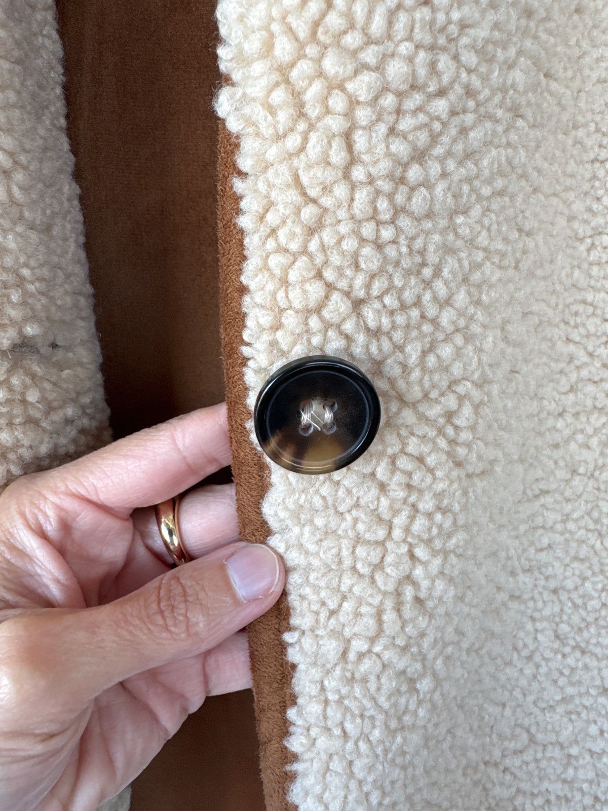 Close-up of a cream sherpa fleece jacket with a brown suede-like trim and a dark button, held by a hand. A beige sherpa fleece button-up coat with brown trim and a brown inner lining, hanging on a metal hanger against a light blue wall. Zara teddy bear jacket, Zara teddy jacket, women’s teddy jacket, teddy fleece jacket, sherpa jacket, button-down jacket, cozy women’s jacket, plush jacket, fall jacket, winter jacket, Edmonton consignment, women’s clothing consignment