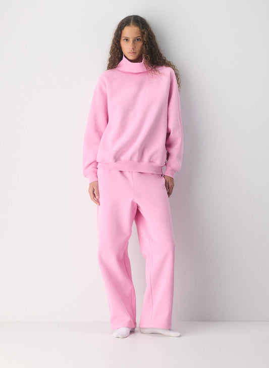 A woman stands against a plain light gray wall wearing a matching light pink lounge set consisting of a mock-neck sweatshirt and barrel sweat pants from Aritzia, visit The RAK to buy them for less, in Sherwood Park