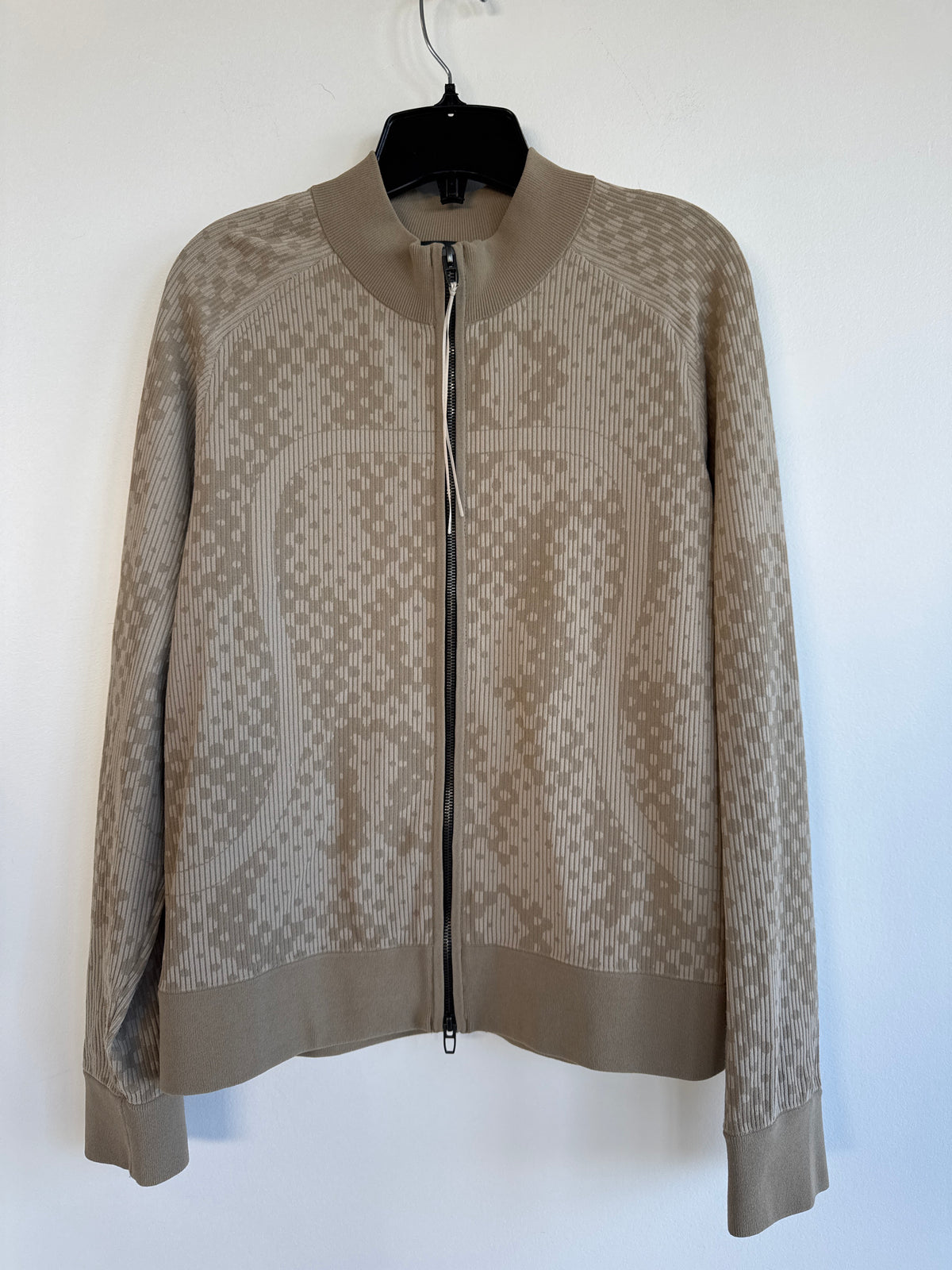 A beige and taupe rare lululemon knit zip-up jacket with a textured geometric pattern, hanging on a black hanger against a plain white wall. Sherwood Park consignment, Sherwood Park shopping, consignment store Sherwood Park, Edmonton consignment, secondhand shopping, secondhand clothing, women’s consignment, women’s clothing consignment, curated consignment, quality secondhand clothing, pre-loved clothing, thrift shopping, sustainable fashion, local shopping, Alberta consignment