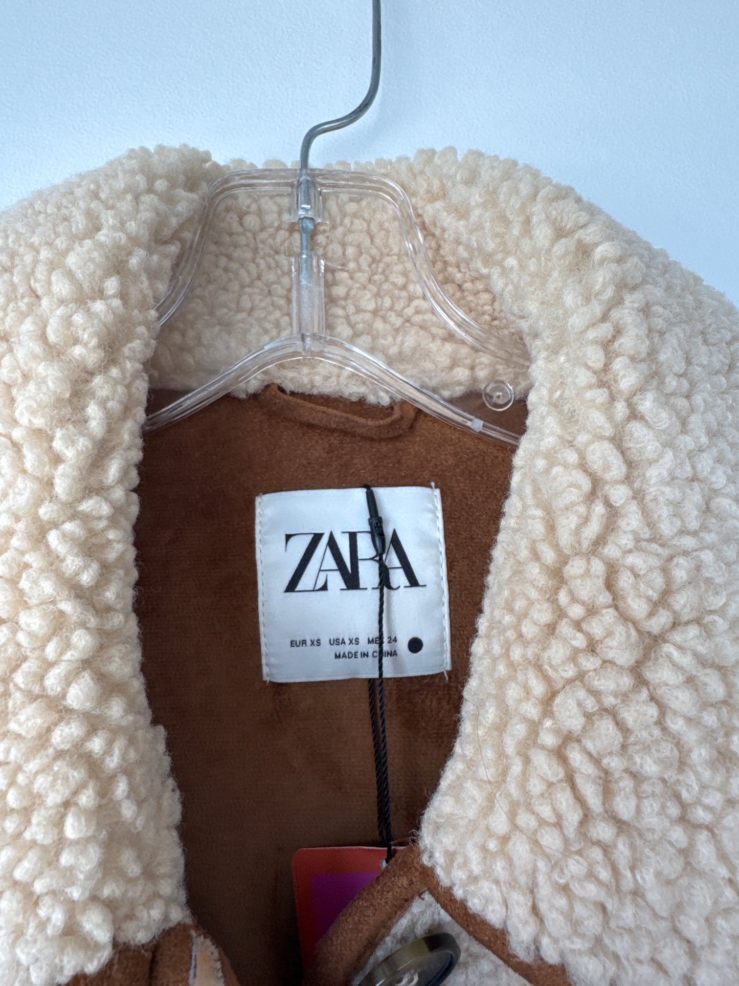 A close-up of a ZARA shearling jacket hanging on a clear plastic hanger against a light blue background. The jacket features a thick, textured cream-colored sherpa collar and a brown inner lining, with the white ZARA brand label clearly visible in the center. A beige sherpa fleece button-up coat with brown trim and a brown inner lining, hanging on a metal hanger against a light blue wall. Zara teddy bear jacket, Zara teddy jacket, women’s teddy jacket, teddy fleece jacket, sherpa jacket, button-down jacket,