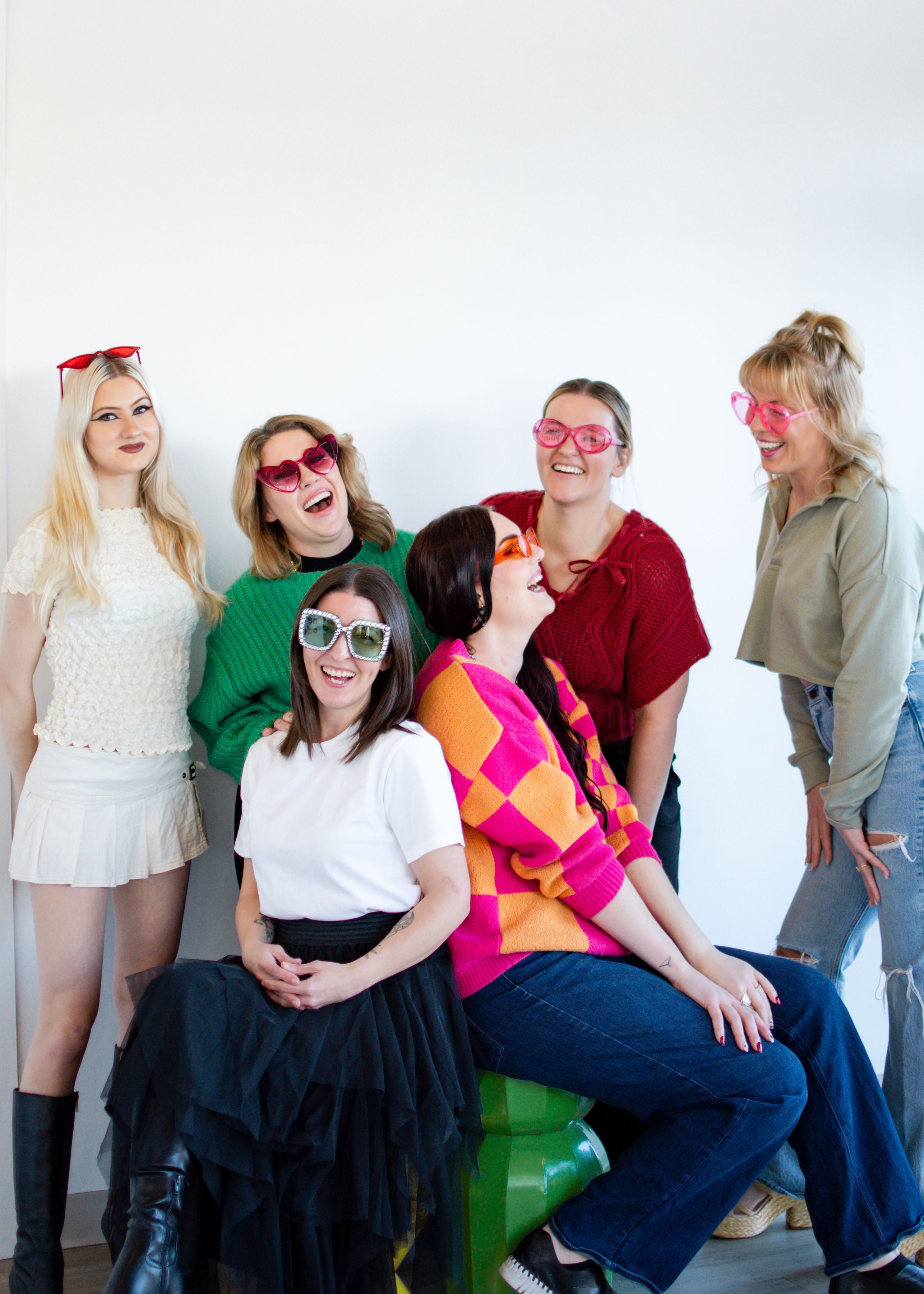 Group of women wearing colorful sunglasses and posing together against a white wall.