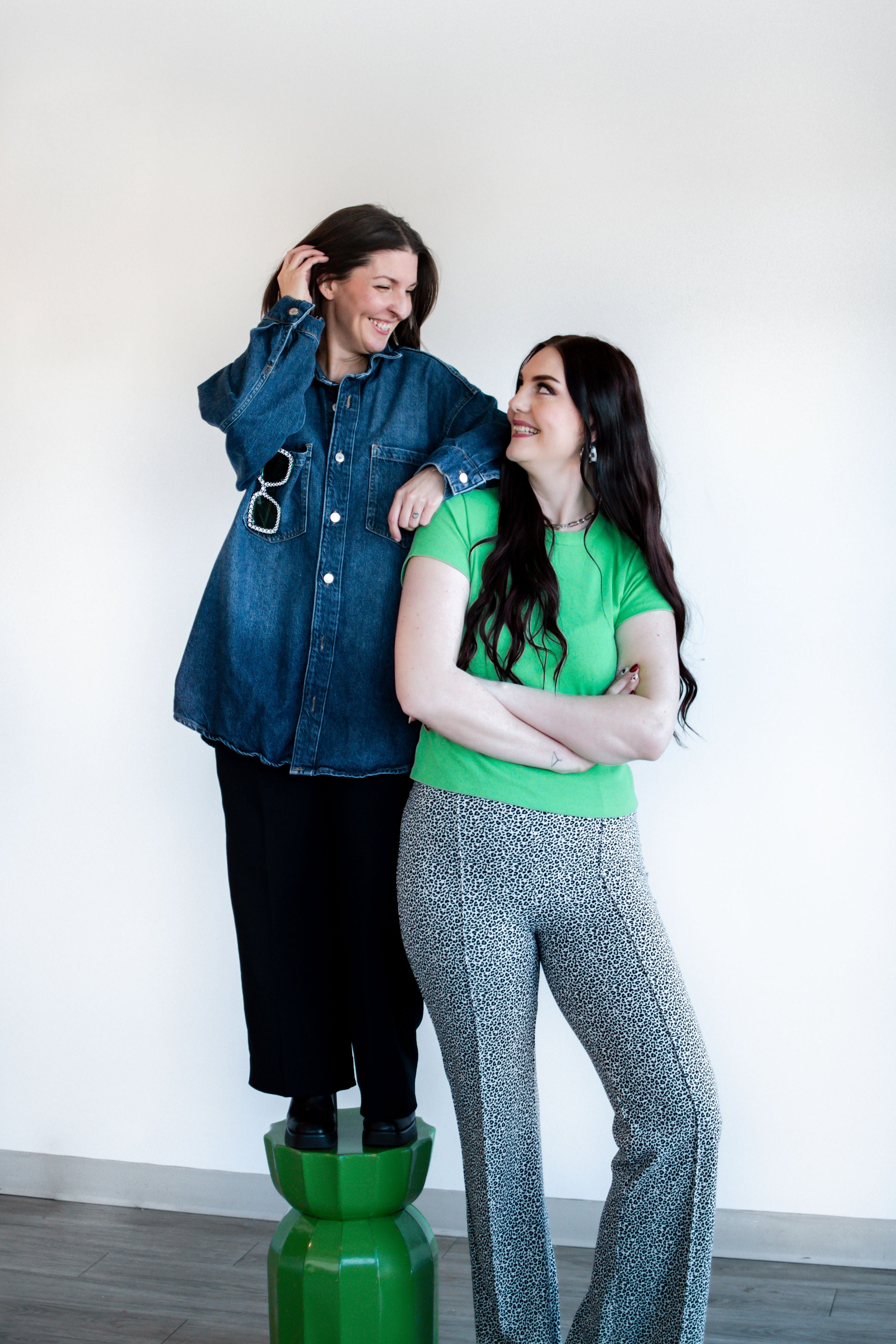Two women standing together, one in a denim jacket and black pants, the other in a green shirt and gray pants, against a white wall.