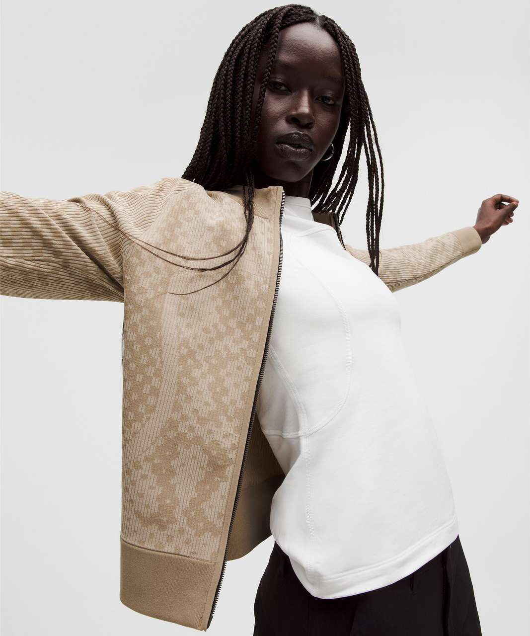 A model wearing a beige and brown patterned knit zip-up jacket over a white long-sleeve top and black pants, posed against a light gray studio background. Rare Lululrmon zip up. Sherwood Park consignment, Sherwood Park shopping, consignment store Sherwood Park, Edmonton consignment, secondhand shopping, secondhand clothing, women’s consignment, women’s clothing consignment, curated consignment, quality secondhand clothing, pre-loved clothing, thrift shopping, sustainable fashion, local shopping, Alberta con