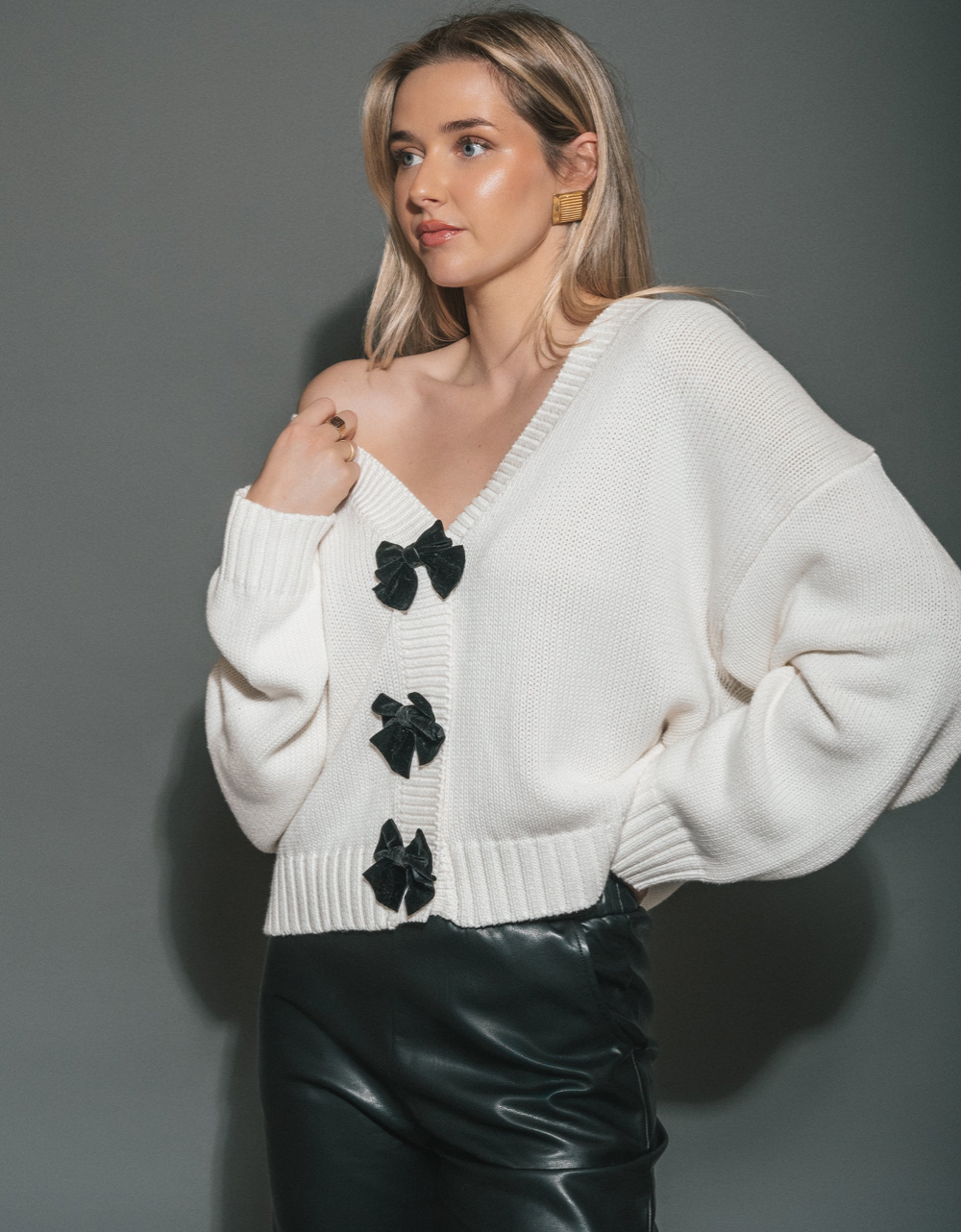 A woman wearing a white ribbed knit cardigan with black bow accents down the front, paired with black faux-leather pants, posed against a solid gray studio background. SHPK consignment, Sherwood Park shop local. Brunette The Label cheaper