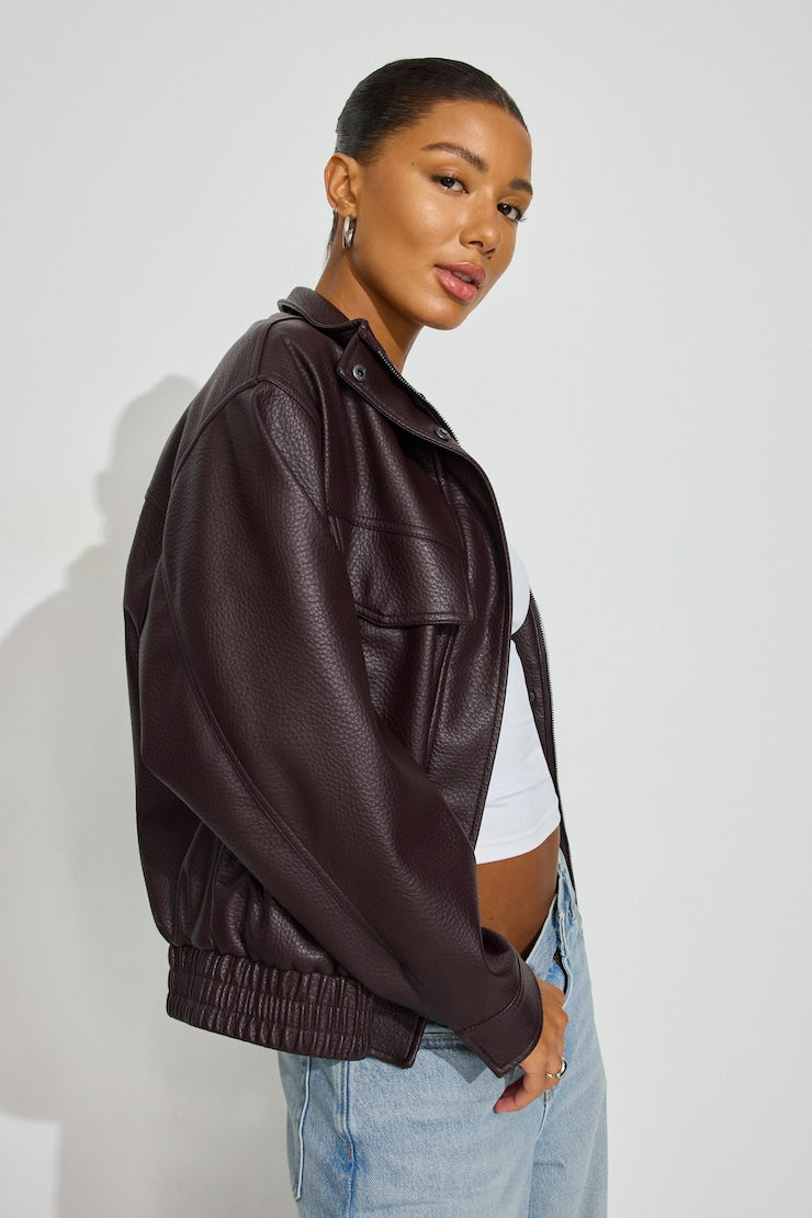 A woman wearing a dark brown faux-leather jacket, styled with a white crop top and light blue jeans, photographed against a plain light gray studio background. Sherwood Park consignment, Sherwood Park shopping, consignment store Sherwood Park, Edmonton consignment, secondhand shopping, secondhand clothing, women’s consignment, women’s clothing consignment, curated consignment, quality secondhand clothing, pre-loved clothing, thrift shopping, sustainable fashion, local shopping, Alberta consignment