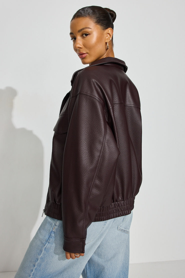 A woman wearing a dark wine Garage faux-leather jacket with a high collar and elasticated hem, paired with light blue jeans, shown from a side-back angle against a light gray studio background. women’s clothing consignment, curated consignment, quality secondhand clothing, pre-loved clothing, thrift shopping, sustainable fashion, local shop